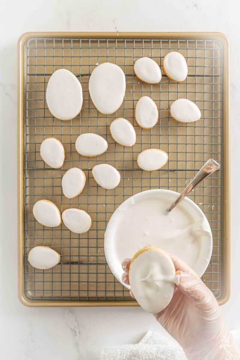 A gloved hand dips an oval cookie into a bowl of white icing, with iced oval cookies drying on a cooling rack in the background.