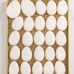 A baking tray with 24 egg-shaped cookies covered in smooth white icing, arranged in neat rows on a cooling rack.