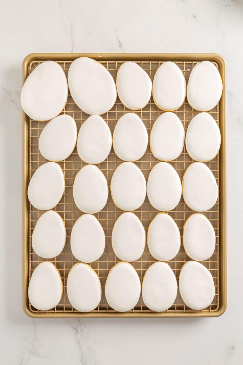 A baking tray with 24 egg-shaped cookies covered in smooth white icing, arranged in neat rows on a cooling rack.