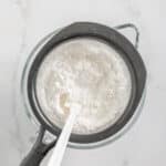 A glass bowl with a fine mesh sieve holding sifted flour, and a white spatula resting inside, on a white marble surface.