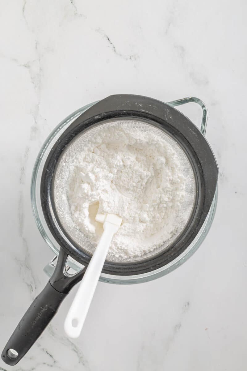 A glass bowl with a fine mesh sieve holding sifted flour, and a white spatula resting inside, on a white marble surface.