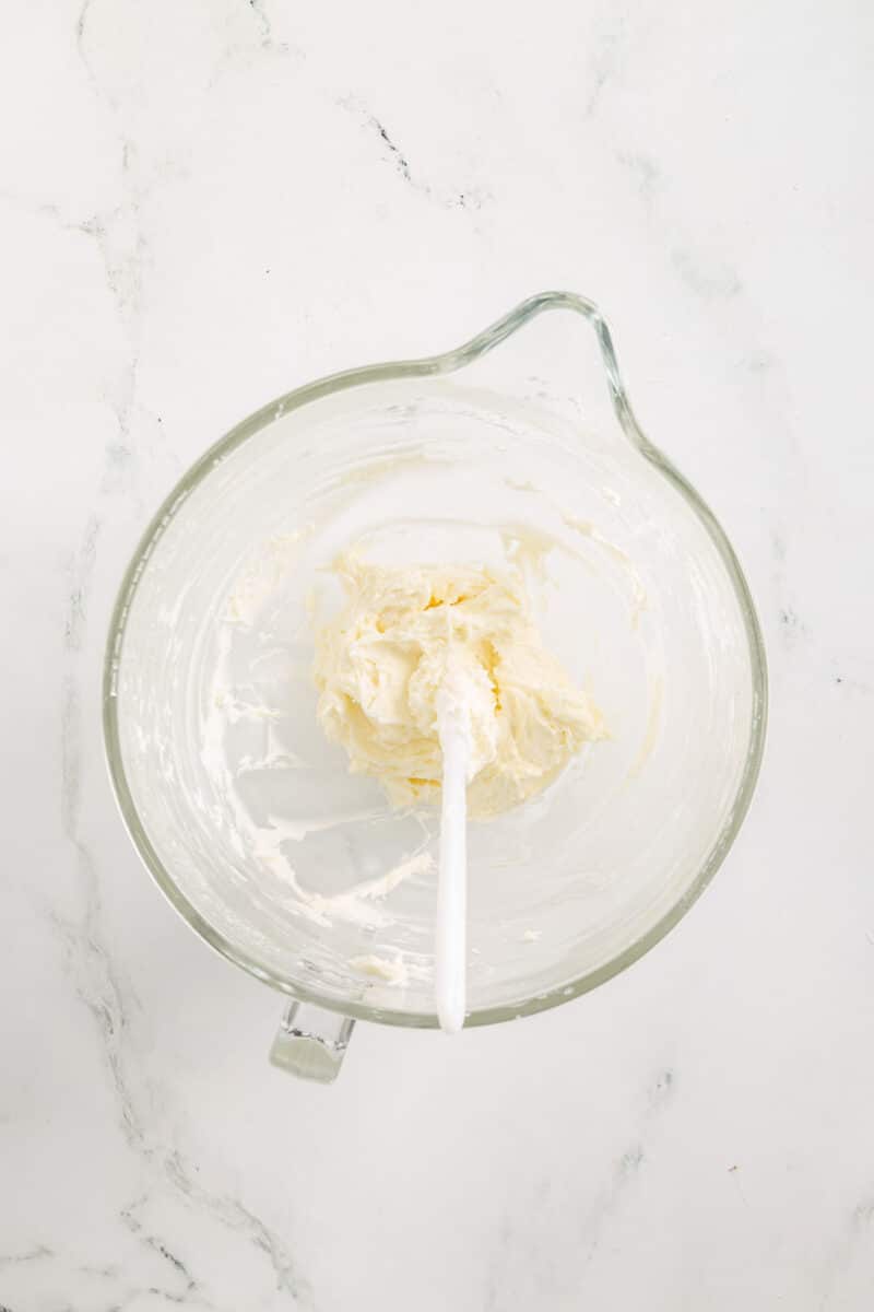 A glass mixing bowl containing a small amount of creamed mixture with a white spatula resting inside, on a white marble surface.