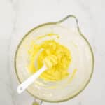 A glass mixing bowl with yellow frosting and a white spatula, with a tube of yellow food coloring lying nearby on a white marble surface.