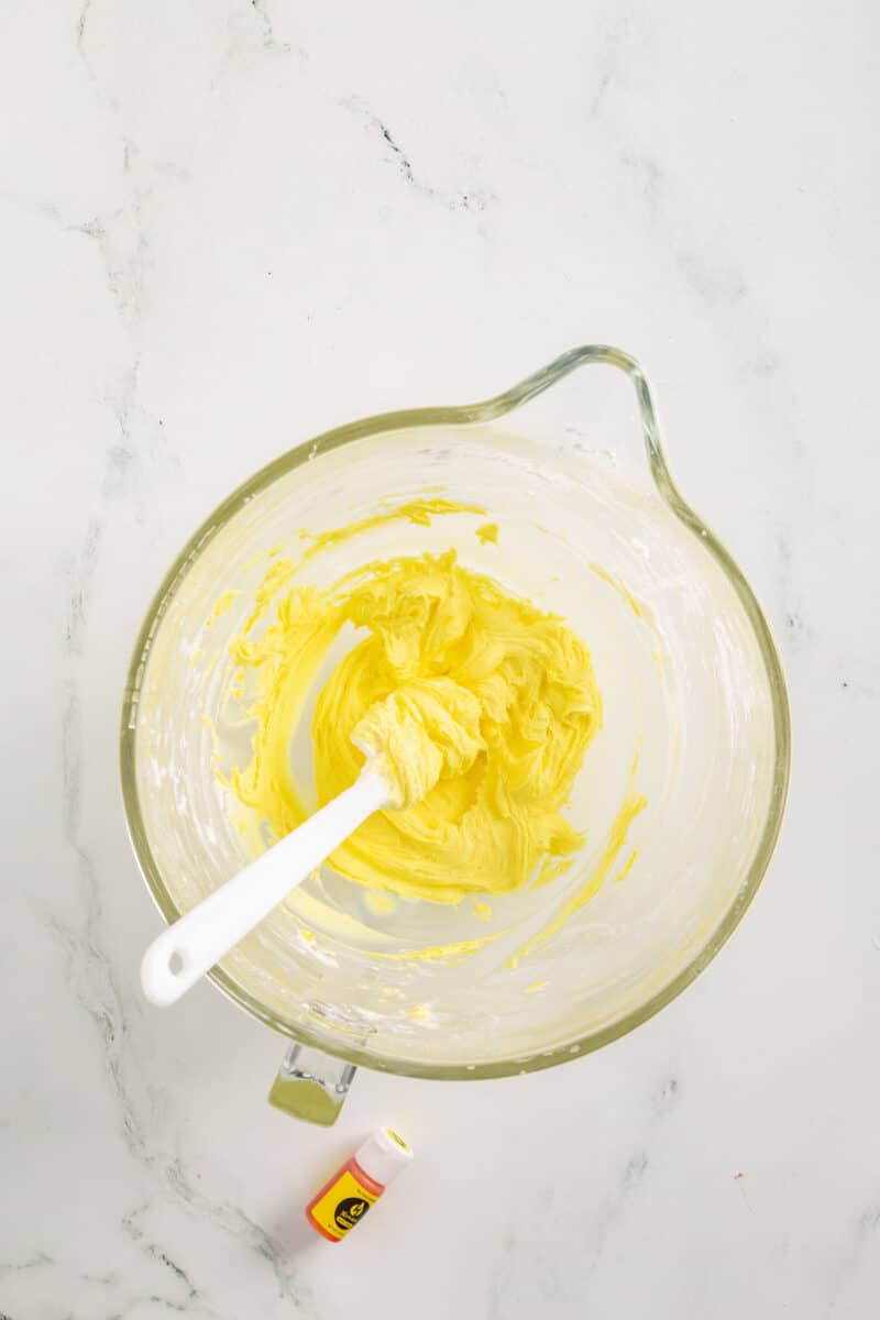 A glass mixing bowl with yellow frosting and a white spatula, with a tube of yellow food coloring lying nearby on a white marble surface.