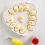 A white decorative plate holds twelve egg-shaped cookies with white icing and yellow frosting; a piping bag with yellow icing and a jar of red sprinkles are nearby.