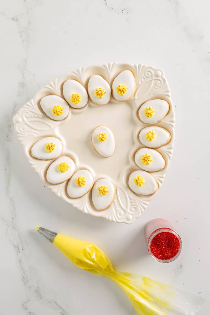 A white decorative plate holds twelve egg-shaped cookies with white icing and yellow frosting; a piping bag with yellow icing and a jar of red sprinkles are nearby.
