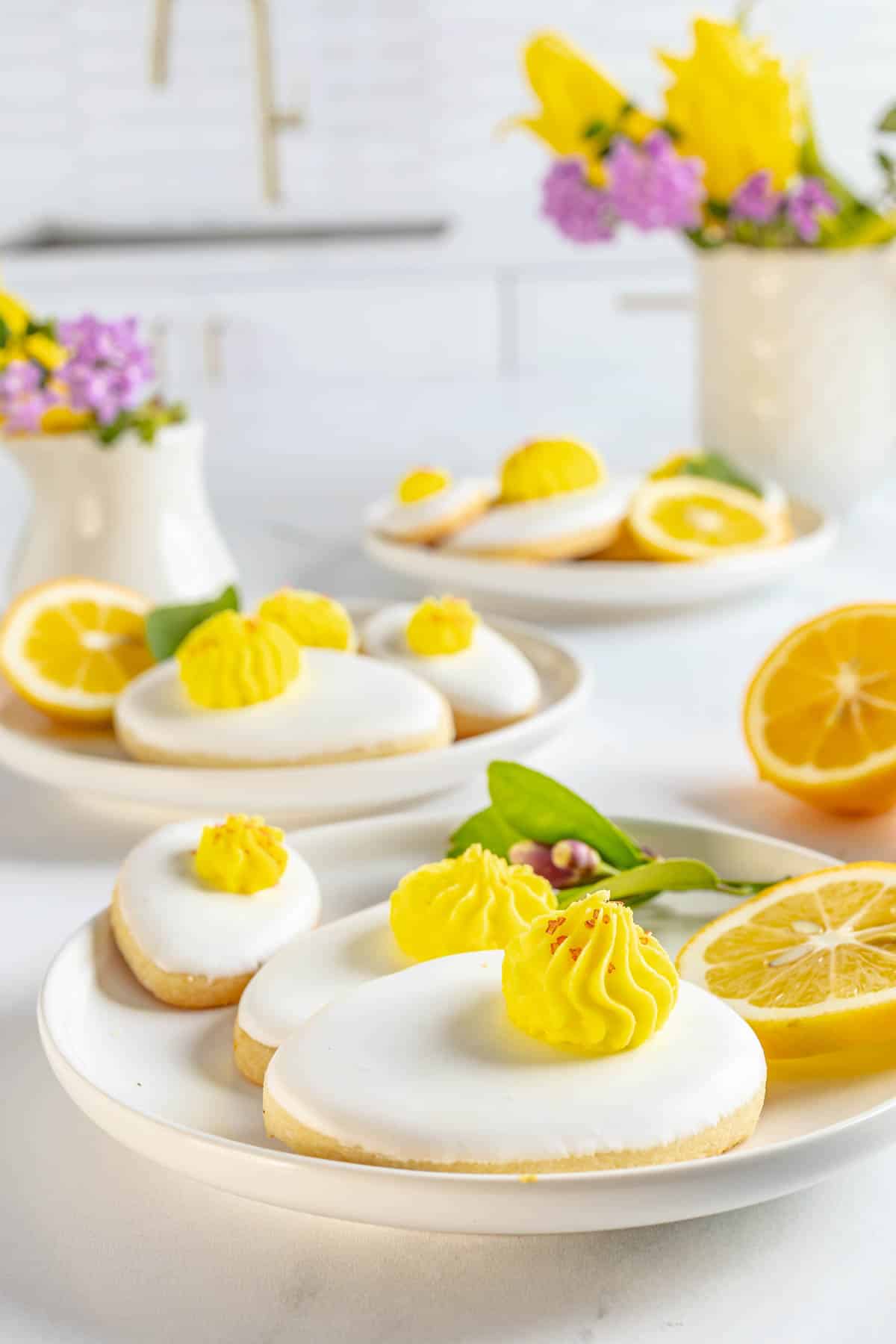 Iced cookies with white frosting and yellow piped topping are arranged on plates, accompanied by sliced lemons and flower decorations on a white countertop.