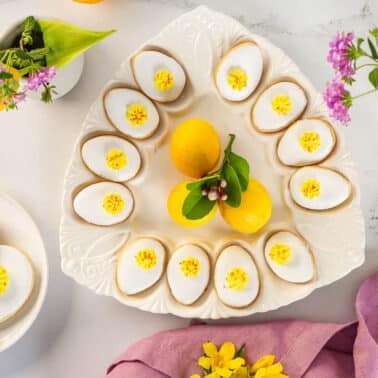A white plate with deviled eggs garnished with yellow filling, surrounded by lemons, flowers, and a pink napkin on a white surface.