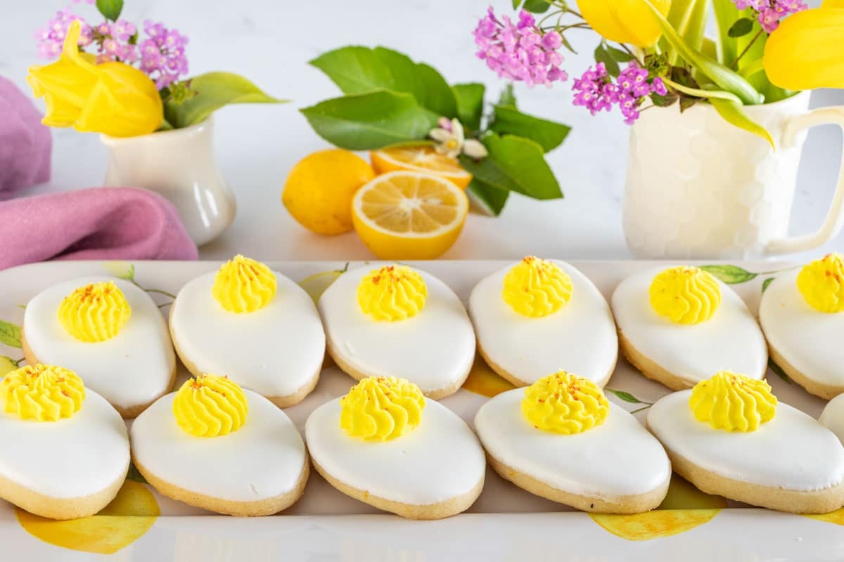 Iced cookies topped with yellow frosting are arranged on a rectangular plate, with yellow flowers and sliced lemons in the background.