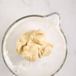 A glass mixing bowl containing a ball of raw cookie dough on a white marble surface.