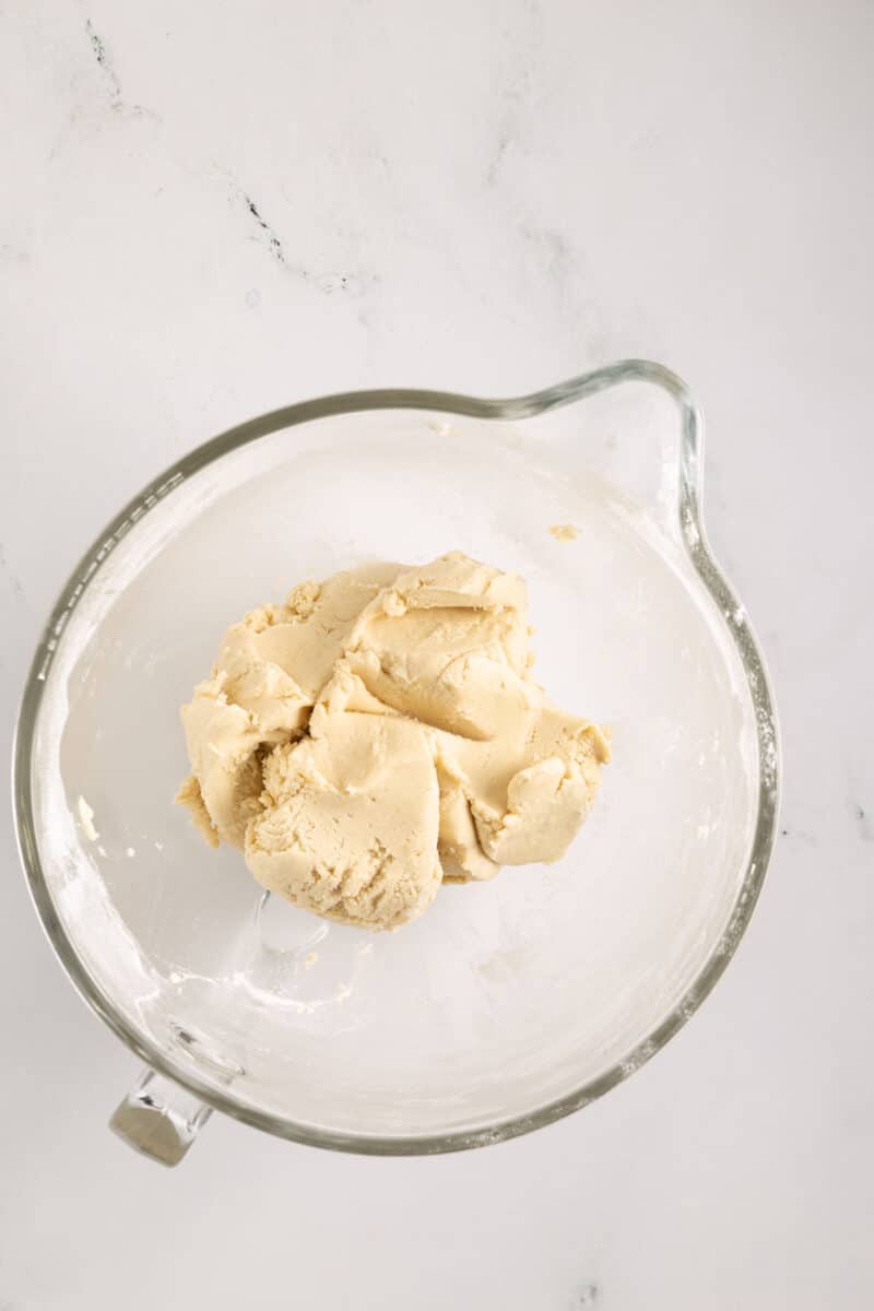 A glass mixing bowl containing a ball of raw cookie dough on a white marble surface.