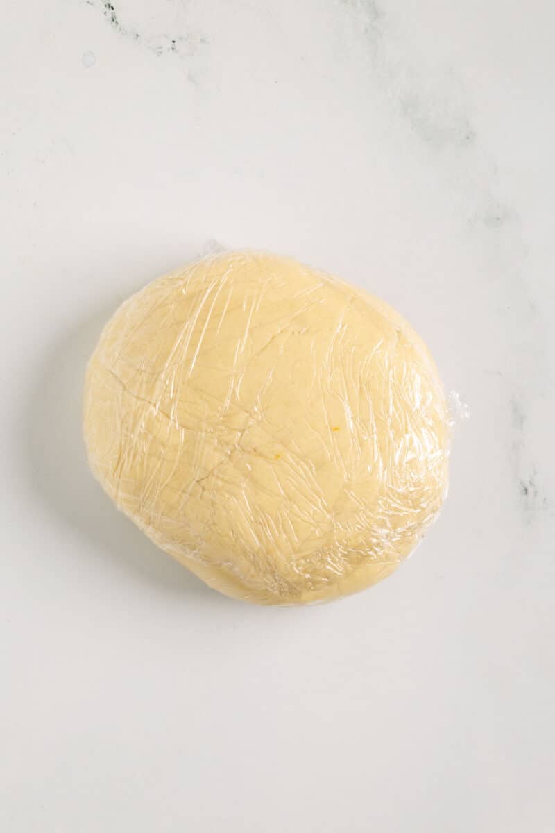 A ball of dough wrapped in plastic wrap on a white marble surface.