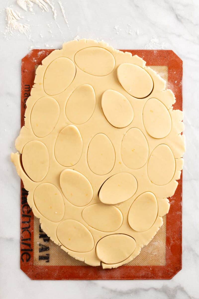 Rolled out cookie dough with oval shapes cut out, placed on a silicone baking mat on a marble surface.