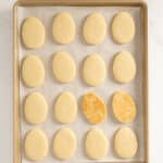 A baking tray with 16 egg-shaped cookies on parchment paper; one cookie has a golden brown surface, while the others are pale.
