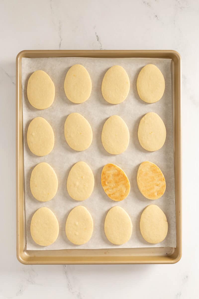A baking tray with 16 egg-shaped cookies on parchment paper; one cookie has a golden brown surface, while the others are pale.
