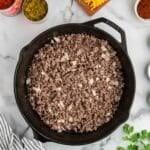 A cast iron skillet with cooked ground beef and onions sits on a marble countertop surrounded by grated cheese, macaroni, canned tomatoes, chopped chilies, and spices.