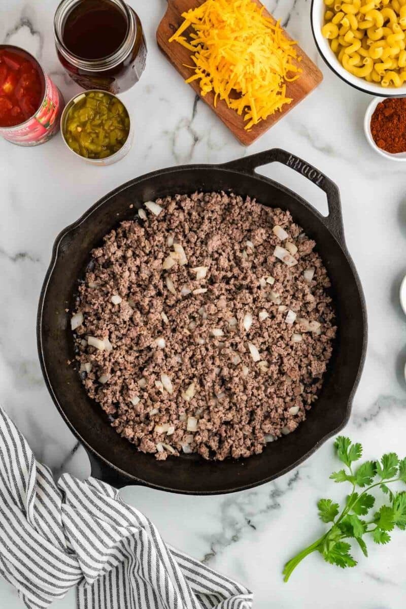 A cast iron skillet with cooked ground beef and onions sits on a marble countertop surrounded by grated cheese, macaroni, canned tomatoes, chopped chilies, and spices.