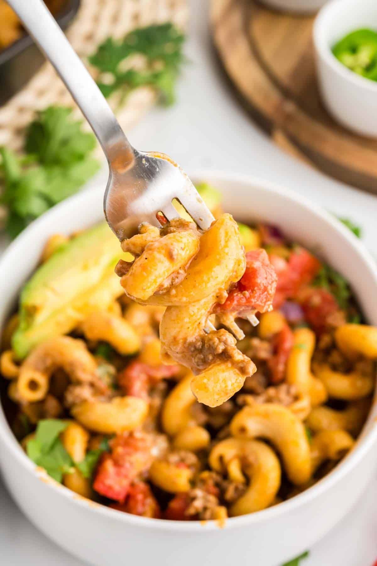 A close-up of a fork holding taco pasta, ground meat, and vegetables above a bowl filled with the same pasta dish.
