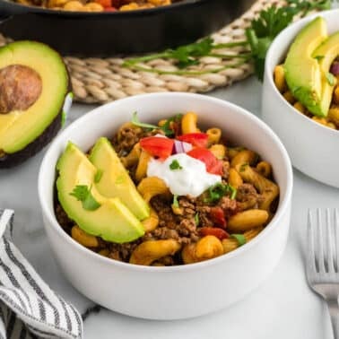 A bowl of taco pasta with ground meat, tomato, sour cream, avocado slices, red onion, and cilantro. Another similar bowl, half an avocado, and two forks are nearby.