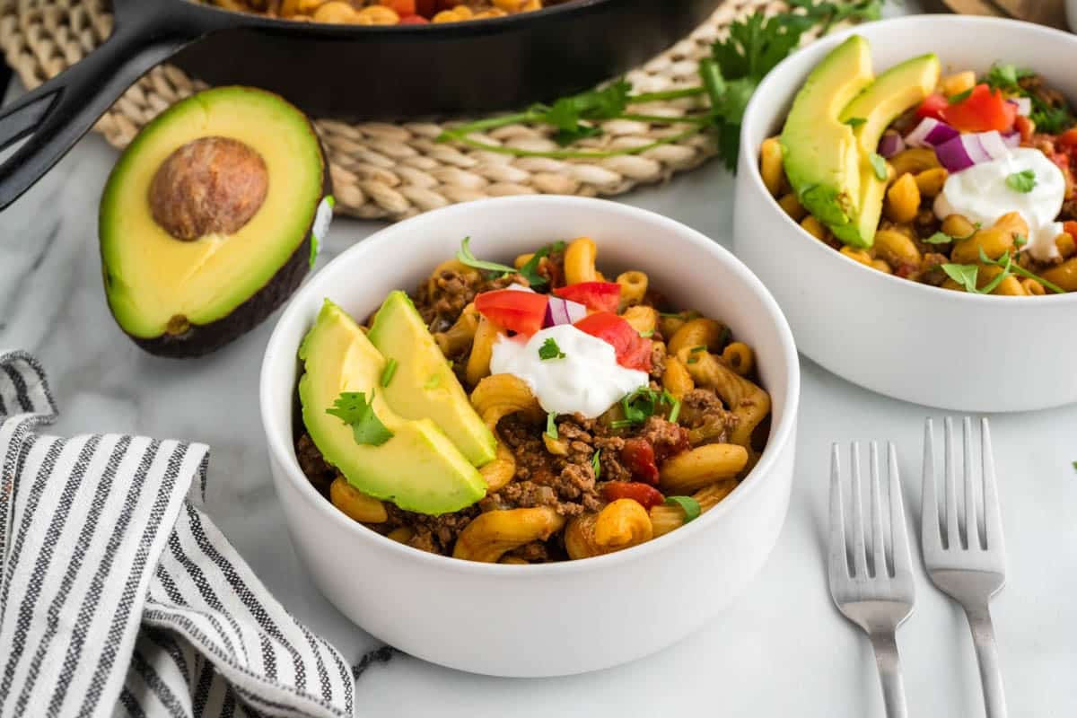 A bowl of taco pasta with ground meat, tomato, sour cream, avocado slices, red onion, and cilantro. Another similar bowl, half an avocado, and two forks are nearby.