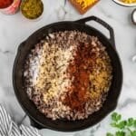 Cast iron skillet with cooked ground beef, onions, and seasonings on a marble counter, surrounded by shredded cheese, macaroni, canned tomatoes, green chiles, and a striped towel.