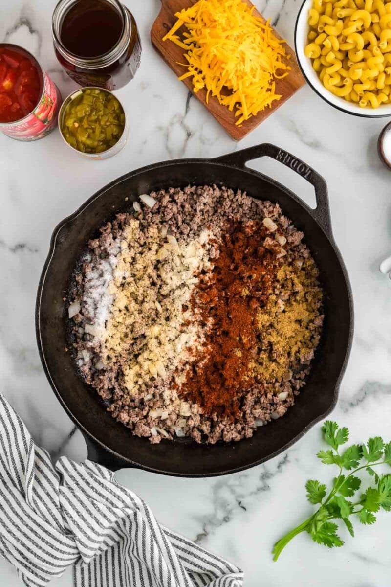 Cast iron skillet with cooked ground beef, onions, and seasonings on a marble counter, surrounded by shredded cheese, macaroni, canned tomatoes, green chiles, and a striped towel.