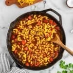 A skillet filled with macaroni, ground beef, tomatoes, and sauce sits on a marble surface with a wooden spoon, grated cheese, and cilantro nearby.