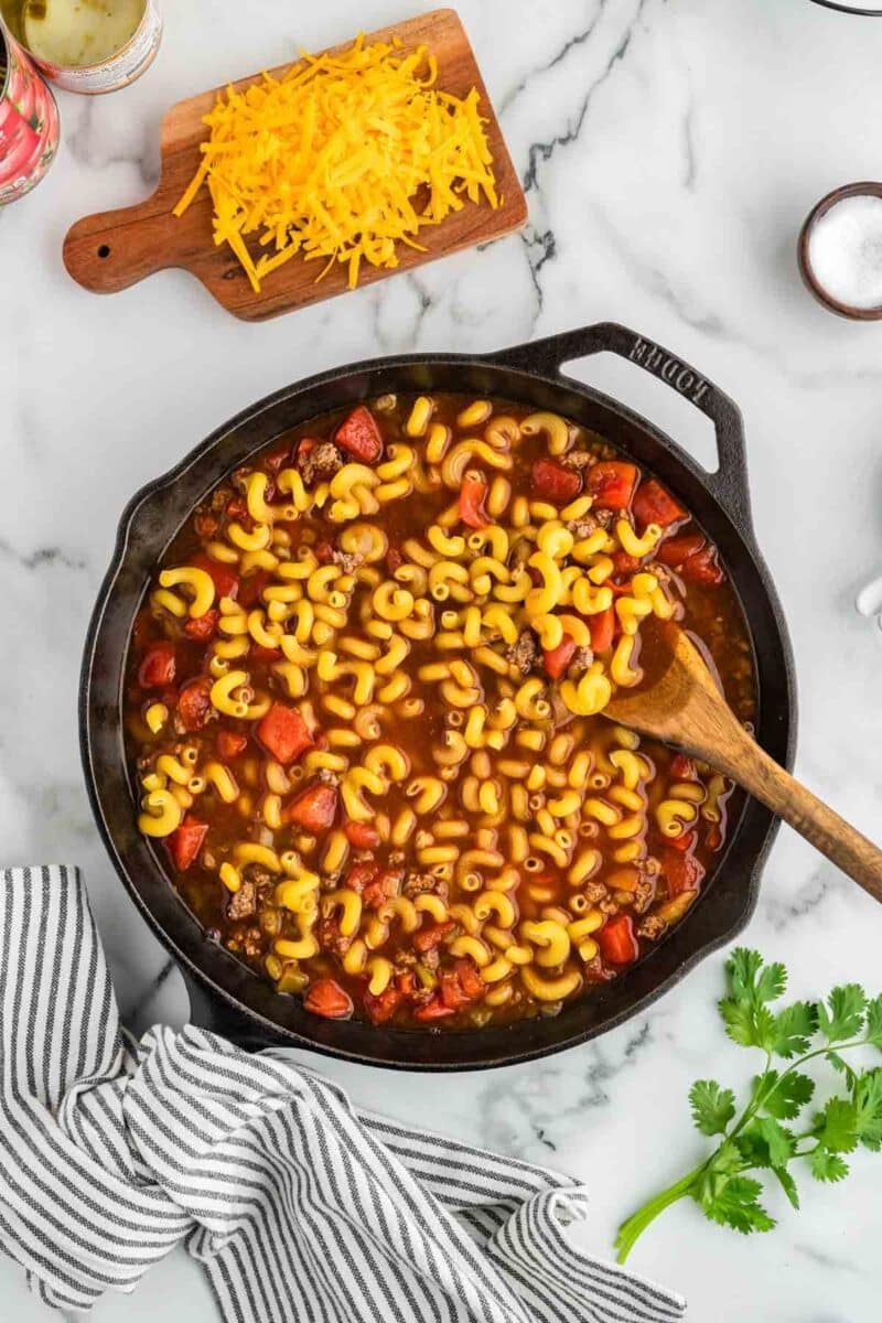 A skillet filled with macaroni, ground beef, tomatoes, and sauce sits on a marble surface with a wooden spoon, grated cheese, and cilantro nearby.