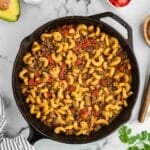 A cast iron skillet filled with cooked pasta, ground meat, and diced tomatoes is surrounded by bowls of sour cream, chopped onions, cilantro, tomato, jalapeño, and an avocado half.