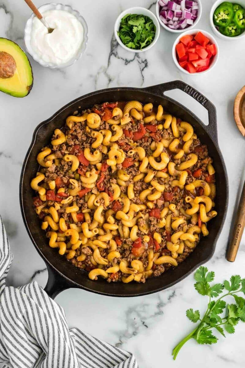 A cast iron skillet filled with cooked pasta, ground meat, and diced tomatoes is surrounded by bowls of sour cream, chopped onions, cilantro, tomato, jalapeño, and an avocado half.