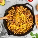 A skillet filled with cheesy pasta mixed with ground beef and vegetables, surrounded by small bowls of chopped cilantro, red onion, tomato, avocado, and sour cream on a marble countertop.