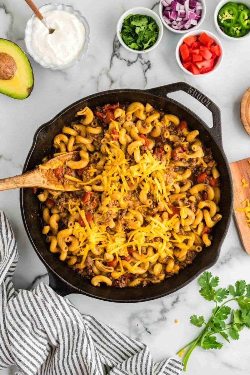A skillet filled with cheesy pasta mixed with ground beef and vegetables, surrounded by small bowls of chopped cilantro, red onion, tomato, avocado, and sour cream on a marble countertop.