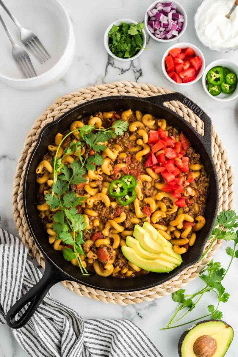 A cast iron skillet with macaroni, ground meat, tomatoes, avocado, cilantro, and jalapeño slices, surrounded by small bowls of chopped toppings on a white countertop.