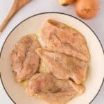 Four raw chicken breasts seasoned with herbs in a white dish next to three whole onions and a wooden spoon on a light surface.