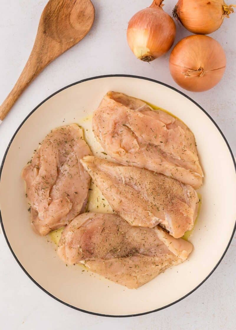 Four raw chicken breasts seasoned with herbs in a white dish next to three whole onions and a wooden spoon on a light surface.
