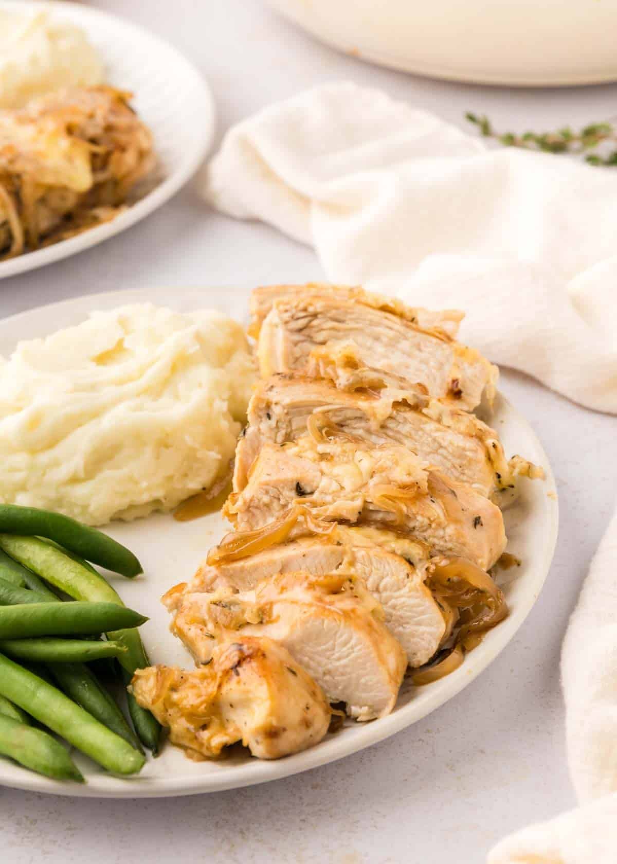 Sliced french onion chicken served with mashed potatoes and green beans on a white plate.
