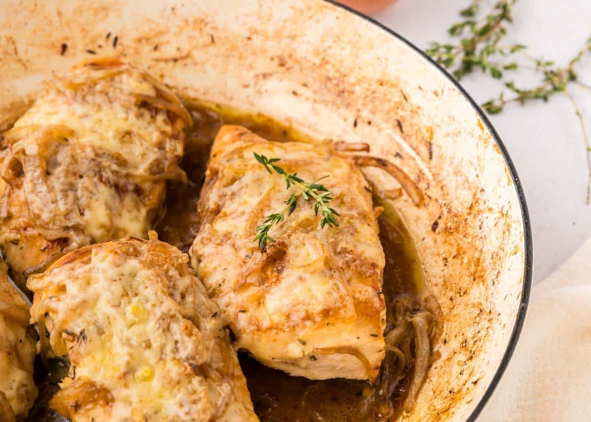 Three pieces of cooked french onion chicken topped with melted cheese and herbs in a white enameled pan, surrounded by caramelized onions and fresh thyme.