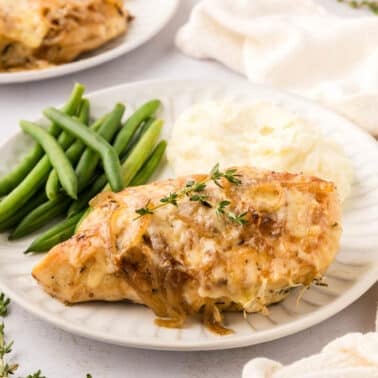 A plate with french onion chickenย topped with herbs and onions, served with green beans and mashed potatoes.