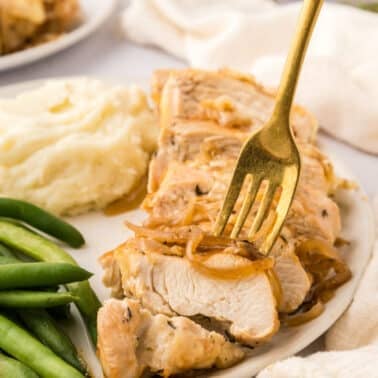 A plate with sliced cooked french onion chicken topped with onions, mashed potatoes, and green beans; a fork is holding a piece of chicken.