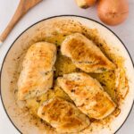 Four cooked, seasoned chicken breasts in a round white skillet, with three whole onions and a wooden spoon beside the pan on a white surface.