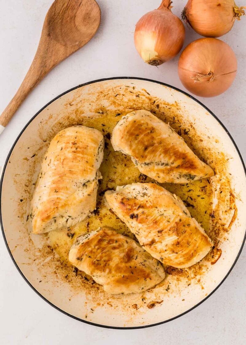 Four cooked, seasoned chicken breasts in a round white skillet, with three whole onions and a wooden spoon beside the pan on a white surface.