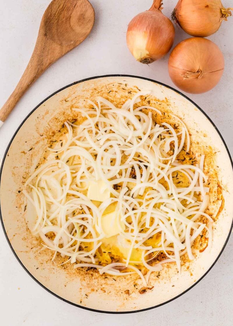 Sliced onions and butter in a white pot, next to three whole onions and a wooden spoon on a white surface.
