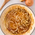 A white pan with sliced onions cooking in brown broth, next to three whole onions and a wooden spoon on a white surface.
