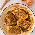 Cooked chicken breasts topped with caramelized onions in a white pan, with whole onions and a wooden spoon nearby.