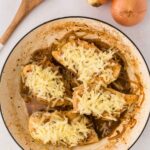 A skillet with chicken breasts topped with shredded cheese and cooked onions, next to whole onions and a wooden spoon on a light surface.