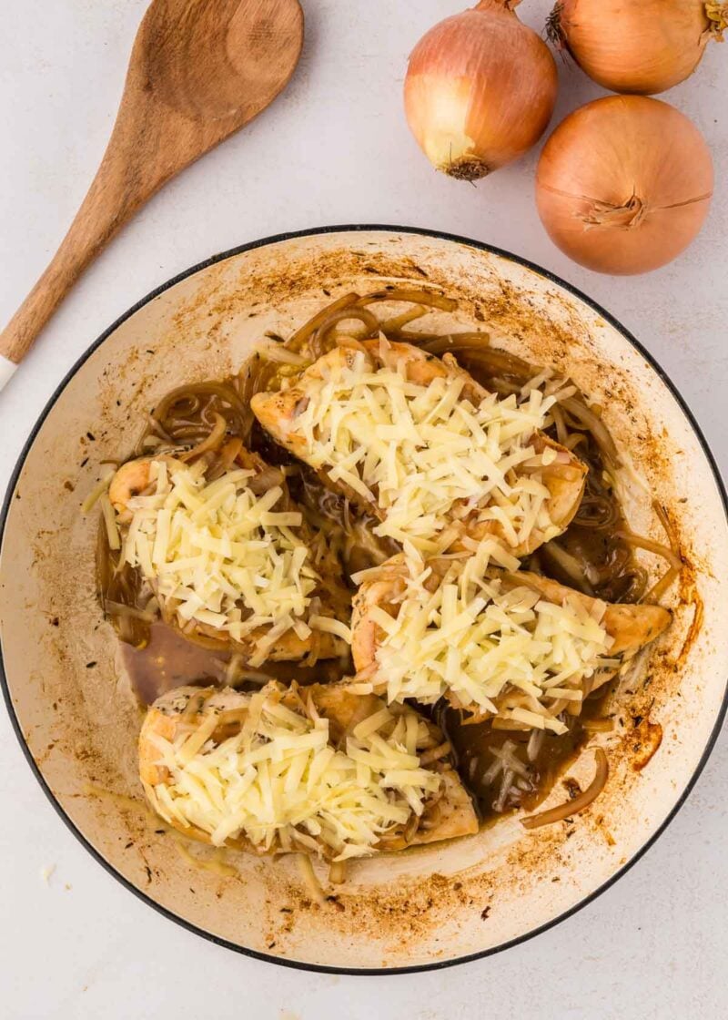 A skillet with chicken breasts topped with shredded cheese and cooked onions, next to whole onions and a wooden spoon on a light surface.
