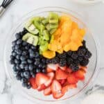 A glass bowl filled with sliced kiwi, mandarin oranges, blackberries, strawberries, and blueberries on a marble surface. A whisk and bowl of dressing are nearby.