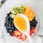 A bowl of assorted fresh fruits including blueberries, blackberries, strawberries, kiwi, and mandarin oranges with a spoonful of orange dressing held above.