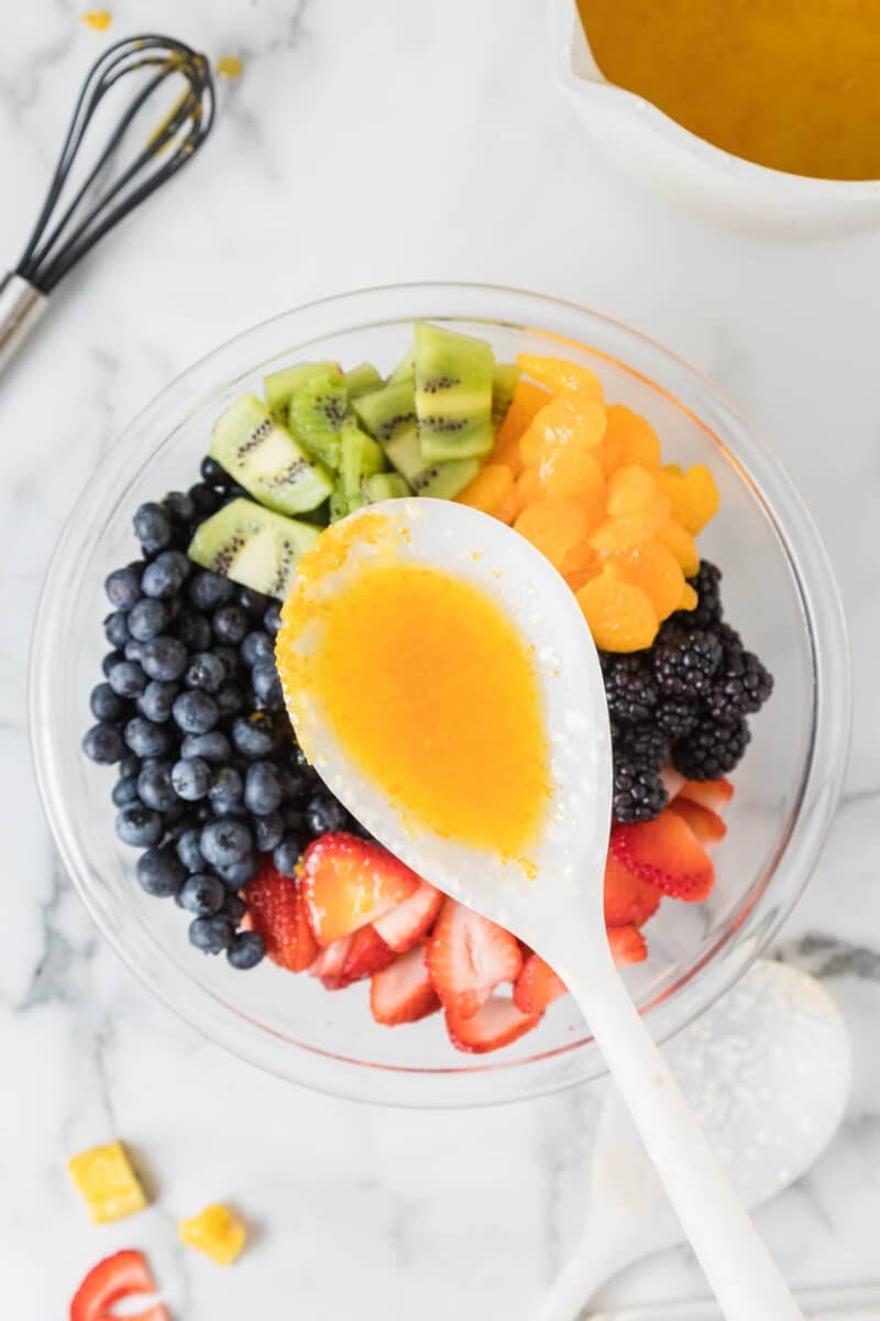 A bowl of assorted fresh fruits including blueberries, blackberries, strawberries, kiwi, and mandarin oranges with a spoonful of orange dressing held above.