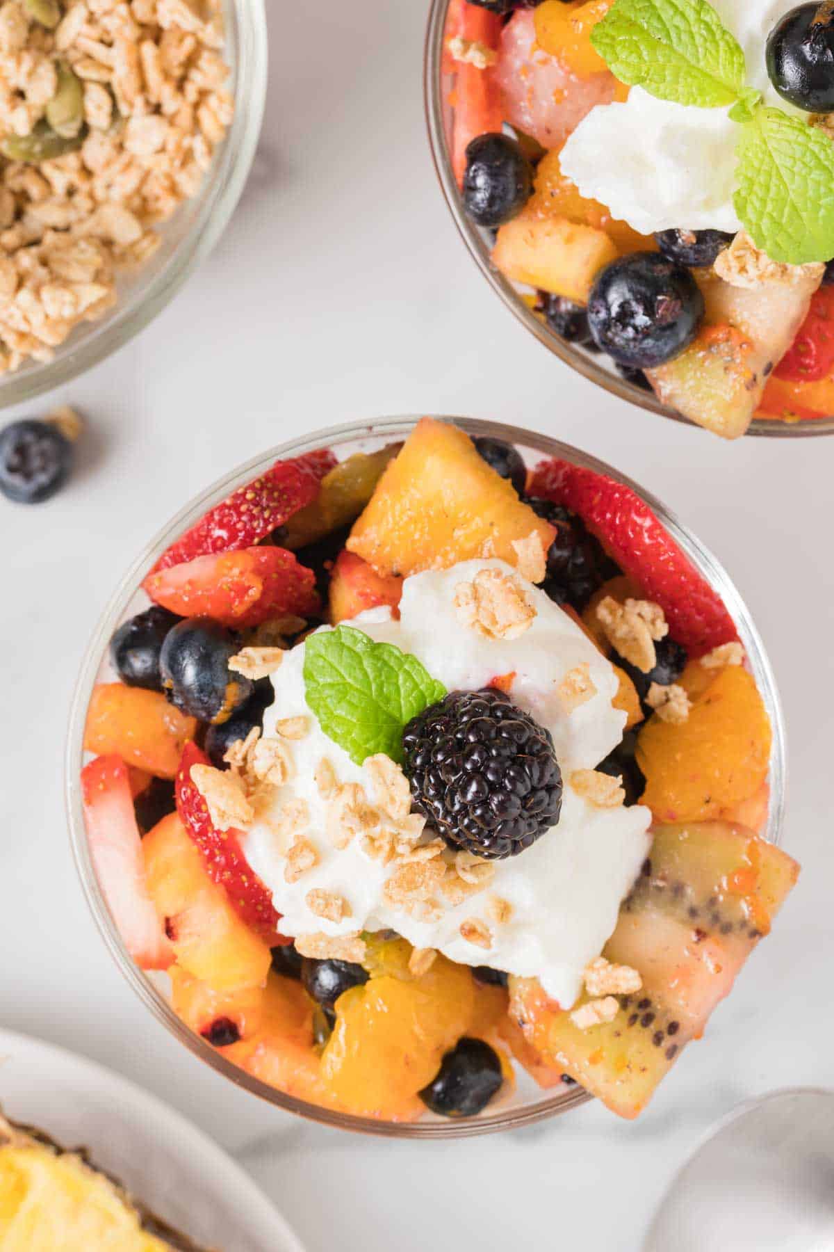 A bowl of mixed fruit salad topped with whipped cream, granola, a blackberry, and a mint leaf, with another bowl and granola in the background.